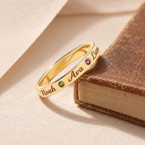Op de afbeelding: Een goudkleurige ring met de gegraveerde namen Noah, Ava en Liam in een cursief lettertype. De ring is versierd met twee kleine, ronde, verzonken edelstenen, een groene en een paarse, die tussen de gegraveerde namen zijn geplaatst.