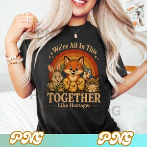 May include: Black t-shirt featuring a colorful graphic of a fox, rabbit, frog, chicks, penguin, and cat under a rainbow. Text reads "We're All In This Together - Like Hostages". The design is whimsical and features cartoon-style animals.