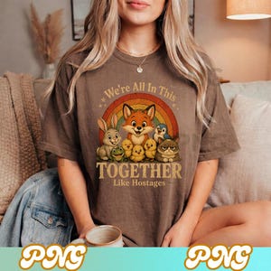 May include: Brown t-shirt with a colorful graphic featuring a fox, rabbit, frog, penguin, chicks, and a grumpy cat in front of a rainbow. Text reads "We're All In This Together Like Hostages."