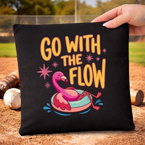 May include: A black square fabric bag featuring a graphic of a pink flamingo floating in a light blue inner tube. The text GO WITH THE FLOW is printed in yellow above the flamingo, accompanied by small pink star-shaped accents and blue water splashes.