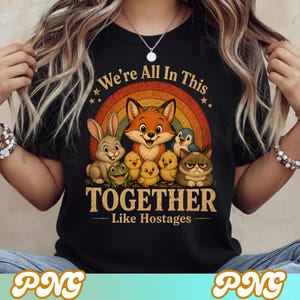 Puede incluir: Camiseta negra con un diseño colorido de un zorro, conejo, rana, pollitos, pingüino y gato frente a un arcoíris. El texto dice "We're All In This Together - Like Hostages". El diseño es caprichoso y de estilo caricaturesco.