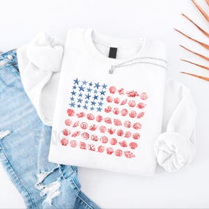 Puede incluir: Una sudadera blanca de manga larga con un gráfico de la bandera estadounidense compuesto por estrellas de mar azules y conchas rojas. La sudadera se combina con unos vaqueros azul claro desgastados y un fino collar de plata sobre el cuello.