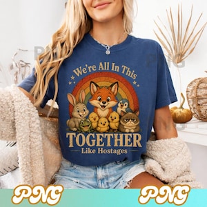 May include: A navy blue t-shirt featuring a rainbow and a group of cartoon animals, including a fox, rabbit, and cat. The text "We're All In This Together Like Hostages" is printed below the animals in a vintage style.
