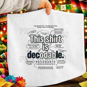 May include: A white t-shirt featuring a black graphic design that explains phonics and wordplay. The text reads: This shirt is decodable, surrounded by arrows and labels detailing digraph explanations, phonics references, and word-decoding instructions.