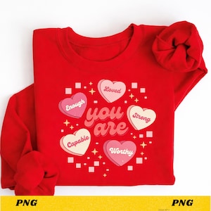 May include: A red sweatshirt with a graphic design featuring the words "you are" in a cursive font, surrounded by heart-shaped candies with positive affirmations like "Enough," "Loved," and "Worthy." The design includes stars and square accents.
