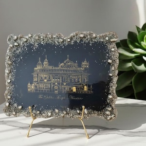 Può includere: Un'opera d'arte decorativa incorniciata con un'illustrazione dorata di un edificio su sfondo blu scuro. La cornice ha un bordo strutturato e scintillante. Il pezzo è esposto su un supporto in metallo dorato.