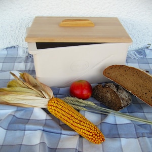 Può includere: Un contenitore per pane in ceramica bianca con un coperchio in legno, una pannocchia di mais e due pagnotte di pane scuro su una tovaglia a quadri blu e bianchi.
