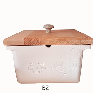 May include: A white ceramic rectangular container with a wooden lid. The lid has a small, round, light-coloured knob. The container has the text "Zwiebel Knoblauch & CO" and a carved onion design. The number "B2" is printed at the bottom.