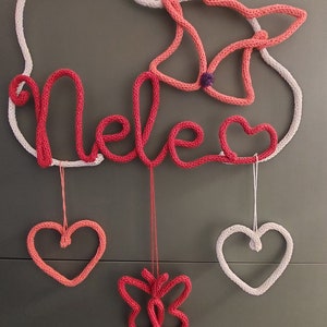 May include: A knitted wall decoration spelling "Nele" in red yarn, with a white cloud outline. It includes a pink butterfly and heart shapes, all hanging by strings. A delightful piece for a child's room or nursery, crafted with care.