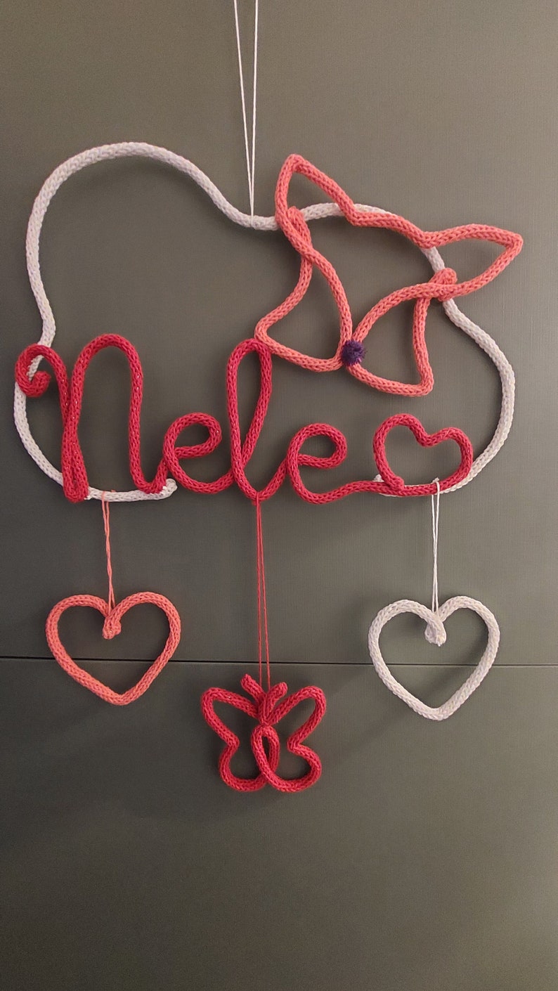 May include: A knitted wall decoration spelling "Nele" in red yarn, with a white cloud outline. It includes a pink butterfly and heart shapes, all hanging by strings. A delightful piece for a child's room or nursery, crafted with care.