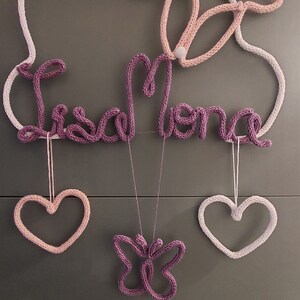 May include: A knitted wall decoration in white, pink, and purple. It features the name "Lisa Mona", a butterfly, and two hearts. The piece is suspended by a white string. The design is suitable for a nursery or child's room.