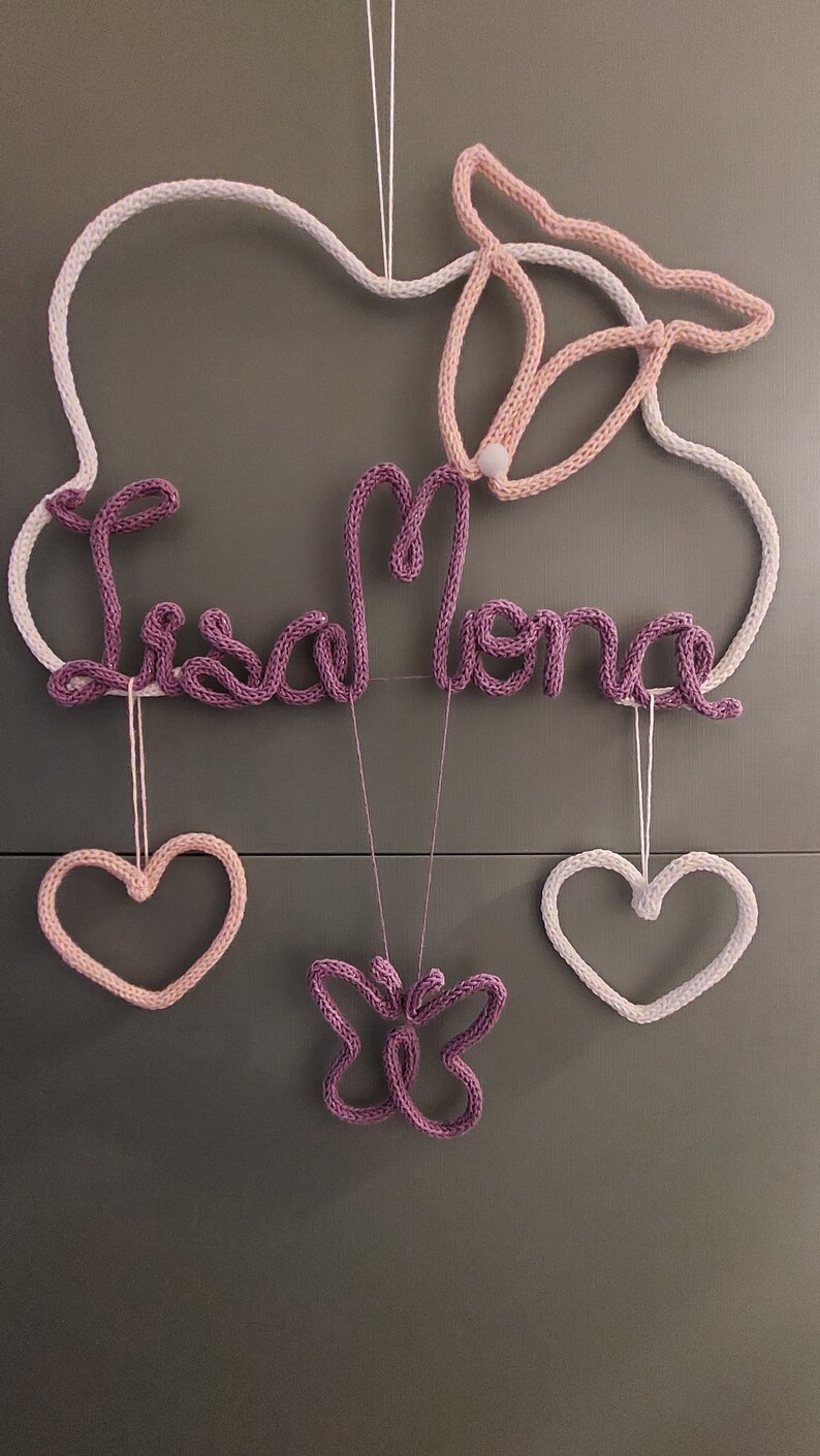 May include: A knitted wall decoration in white, pink, and purple. It features the name "Lisa Mona", a butterfly, and two hearts. The piece is suspended by a white string. The design is suitable for a nursery or child's room.