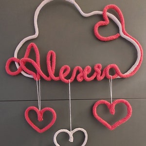 May include: A handmade, pink and white knitted cloud-shaped wall hanging with the name "Alexa" in pink script. Three hanging hearts, two pink and one white, dangle below. A charming nursery or bedroom decoration.