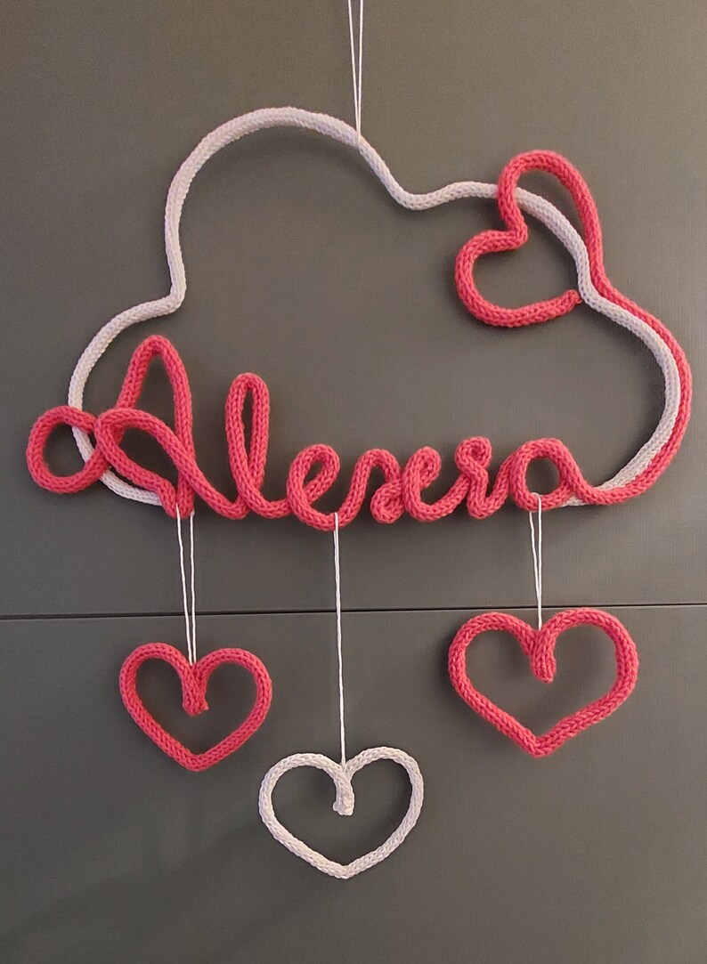 May include: A handmade, pink and white knitted cloud-shaped wall hanging with the name "Alexa" in pink script. Three hanging hearts, two pink and one white, dangle below. A charming nursery or bedroom decoration.