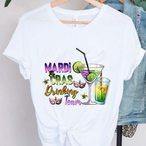 May include: White t-shirt with a Mardi Gras Drinking Team graphic. The graphic features a purple, green, and gold Mardi Gras mask, a cocktail glass with a lime wedge, and a straw. The text "Mardi Gras Drinking Team" is in gold lettering.