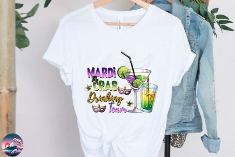 May include: White t-shirt with a Mardi Gras Drinking Team graphic. The graphic features a purple, green, and gold Mardi Gras mask, a cocktail glass with a lime wedge, and a straw. The text "Mardi Gras Drinking Team" is in gold lettering.