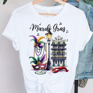 May include: White t-shirt with a colorful Mardi Gras design featuring a mask, beads, a street lamp, a building, and a crawfish. The text "Mardi Gras" is written in black cursive.