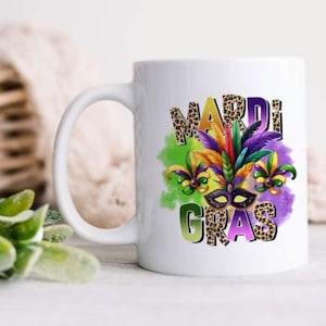 May include: White ceramic coffee mug with a colorful Mardi Gras design. The design features a purple, green, and gold mask with feathers and beads. The words "Mardi Gras" are written in a leopard print font.