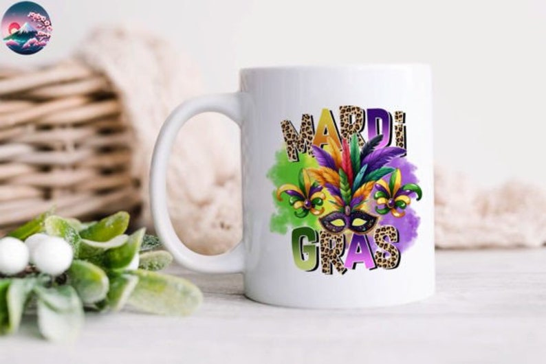 May include: White ceramic coffee mug with a colorful Mardi Gras design. The design features a purple, green, and gold mask with feathers and beads. The words "Mardi Gras" are written in a leopard print font.