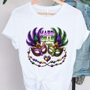 May include: White t-shirt with a Mardi Gras theme. The design features two purple, green, and gold masquerade masks with feathers and beads.