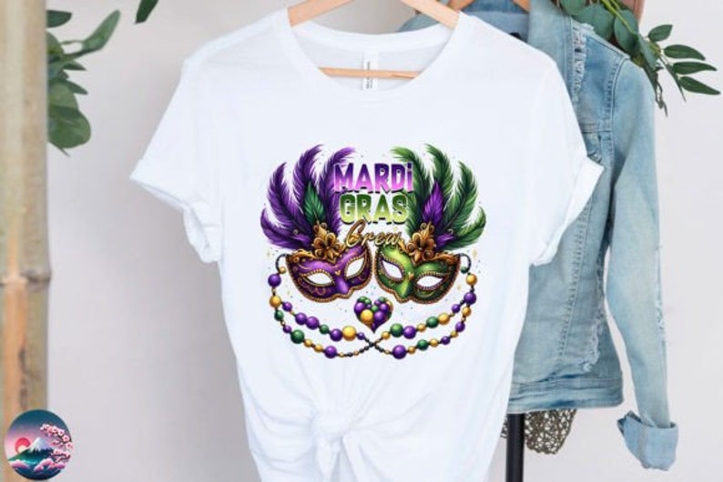 May include: White t-shirt with a Mardi Gras theme. The design features two purple, green, and gold masquerade masks with feathers and beads.