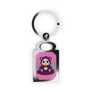 May include: A silver keychain with a rectangular pendant featuring a cartoon bear in a purple hooded outfit, holding a candle. The background is pink with a tombstone and cross.