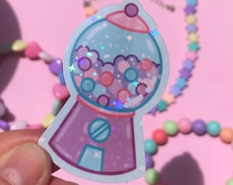 May include: A holographic sticker of a gumball machine in shades of purple, pink, and blue. The gumball machine is filled with colorful gumballs and star-shaped sparkles. The sticker is on a pink background with beaded jewelry.