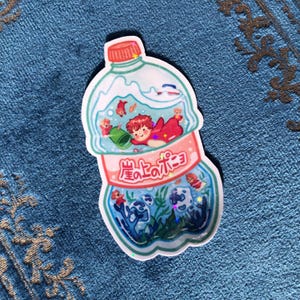 K&ouml;nnte beinhalten: Ein Aufkleber mit einer durchsichtigen Plastikflasche mit einem rosa Etikett, auf dem "星ひとみ" in japanischer Schrift steht. In der Flasche befindet sich eine Comic-Illustration einer Person mit roten Haaren und einem roten Fisch, der in einem blauen und gr&uuml;nen Meer mit Korallen und anderen Fischen schwimmt.