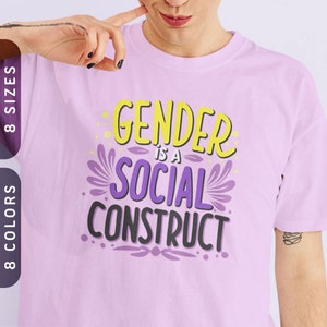 May include: A light purple t-shirt with the text "Gender is a Social Construct" in yellow, purple, and black lettering.