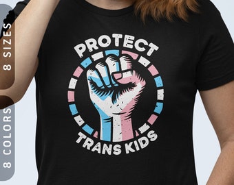 Può includere: Maglietta nera con una grafica di un pugno in un cerchio con le parole "PROTECT TRANS KIDS" intorno. Il pugno è colorato con i colori della bandiera del pride transgender.