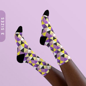 Peut inclure: Une paire de chaussettes avec un motif géométrique de triangles violets, jaunes et noirs. Les chaussettes sont portées sur les jambes d'une personne.