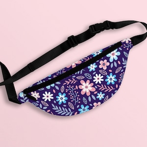 Puede incluir: Una riñonera morada con un estampado floral con flores rosas, azules y blancas. La riñonera tiene una correa negra ajustable.