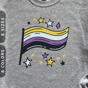 May include: A gray sweatshirt featuring a non-binary pride flag design with yellow, white, purple, and black colors. The design includes stars and a diamond shape. The text "6 SIZES" and "8 COLORS" is on the left side.