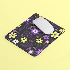 Puede incluir: Una alfombrilla de ratón negra con un patrón floral con flores moradas y amarillas sobre un fondo oscuro. Un ratón de ordenador blanco está encima de la alfombrilla de ratón.