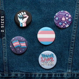May include: Five different button badges with various designs. The badges feature a fist with a transgender flag, a transgender flag, a transgender flag with stars, a floral pattern, and the text "Trans Lives Matter".