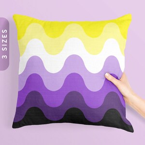 Puede incluir: Un cojín cuadrado con un diseño ondulado en amarillo, blanco, morado y negro. Una mano sostiene el cojín. El texto "3 SIZES" es visible. El diseño es una bandera del orgullo no binario.