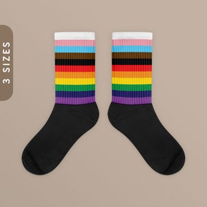 Op de afbeelding: Een paar zwarte sokken met een regenboogstreepjespatroon. De strepen zijn wit, lichtblauw, roze, bruin, oranje, geel, groen en paars. De sokken worden getoond tegen een lichtbruine achtergrond.