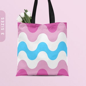 May include: A white tote bag with black handles and a pink and blue wavy pattern. The bag is on a pink background.