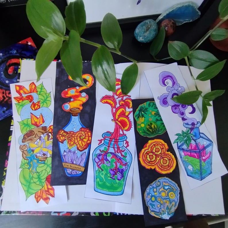 May include: A collection of colorful, hand-drawn bookmarks featuring fantasy-themed illustrations. Designs include potion bottles with crystals, a Venus flytrap, and abstract organic shapes. The bookmarks are on white paper with black backgrounds.