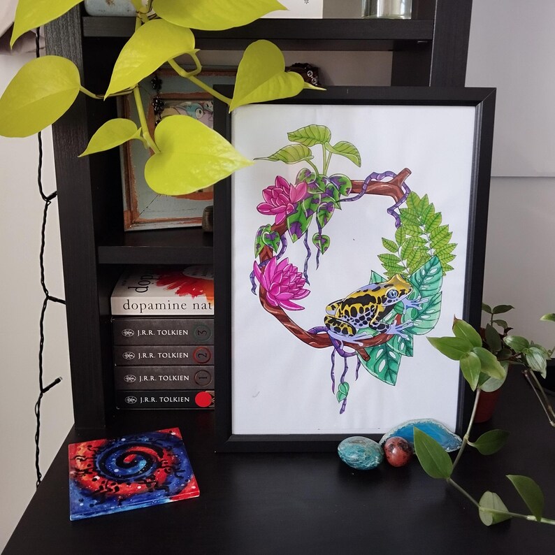 K&ouml;nnte beinhalten: Gerahmter Kunstdruck mit einer farbenfrohen Illustration eines Frosches in einem floralen Kranz. Das Kunstwerk zeigt rosa Lotusbl&uuml;ten, gr&uuml;ne Bl&auml;tter und einen gelb-schwarzen Frosch. Der Rahmen ist schwarz, und der Druck wird auf einer schwarzen Oberfl&auml;che pr&auml;sentiert.
