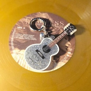 May include: A silver glitter guitar keychain with a black fretboard and white trim rests on a gold vinyl record. The keychain is attached to a silver ring. The record has text that reads "Fearless Taylor's Version".