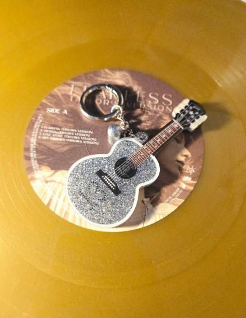 May include: A silver glitter guitar keychain with a black fretboard and white trim rests on a gold vinyl record. The keychain is attached to a silver ring. The record has text that reads "Fearless Taylor's Version".