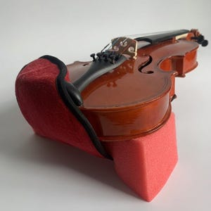 Op de afbeelding: Een gelakte houten viool met een rode schoudersteun en een roze schuimrubberen steun. De viool heeft snaren, een kam en een zwarte toets. De schoudersteun is gemaakt van een zacht, getextureerd materiaal.