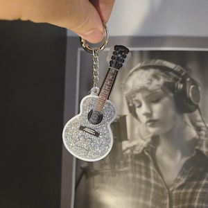 May include: A silver glitter guitar-shaped keychain with a black and brown guitar neck and strings. The keychain is attached to a silver keyring and chain. The background includes a black and white photo.