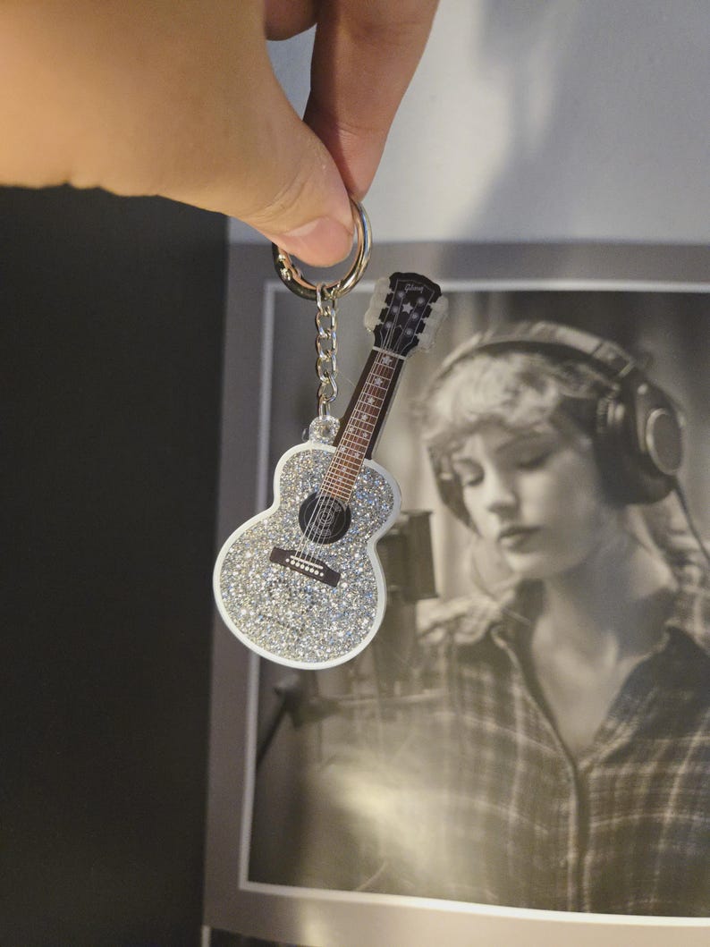 May include: A silver glitter guitar-shaped keychain with a black and brown guitar neck and strings. The keychain is attached to a silver keyring and chain. The background includes a black and white photo.