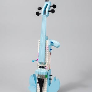 May include: A light blue toy violin made from plastic bricks, with a small figure on top. The word "ARIA" is displayed on the body of the instrument. The violin features black tuning pegs and a black bridge.