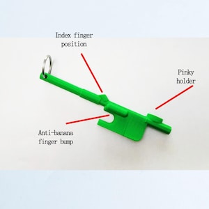 Könnte beinhalten: Ein hellgrünes Kunststoffwerkzeug mit Schlüsselring, das zum Halten einer Banane bestimmt ist. Der Text auf dem Bild lautet: "Index finger position", "Anti-banana finger bump" und "Pinky holder".
