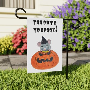 May include: A white Halloween garden flag featuring a cute cartoon mouse wearing a witch's hat and sitting inside a jack-o'-lantern. The text on the flag reads "Too cute to spook!"