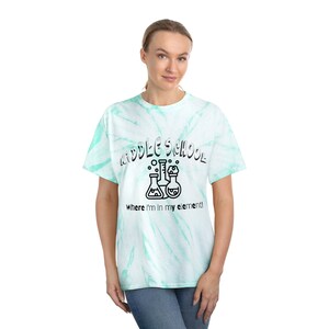 May include: A tie-dye light blue and white t-shirt with the text "Middle School" in a black outline and a graphic of three beakers with bubbles. The text below the graphic says "Where I'm in my element!"