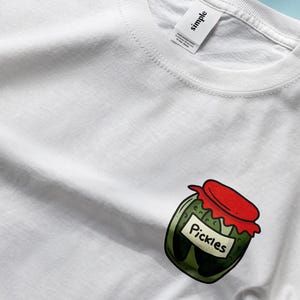 May include: White t-shirt with a cartoon illustration of a jar of pickles. The jar has a red lid and the label says "Pickles".